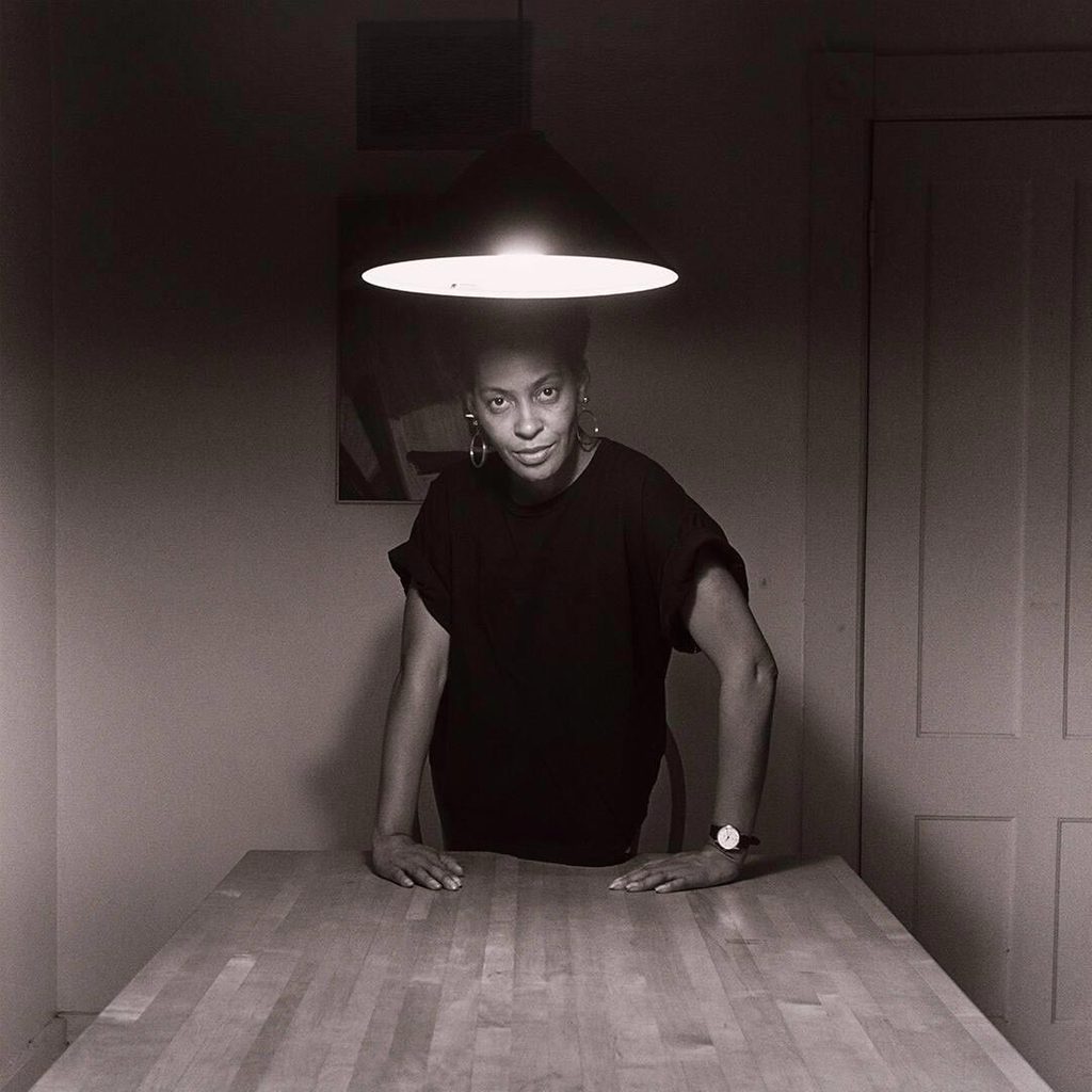 black-and-white photograph of a black woman standing behind a kitchen table underneath a lamp suspended from the ceiling