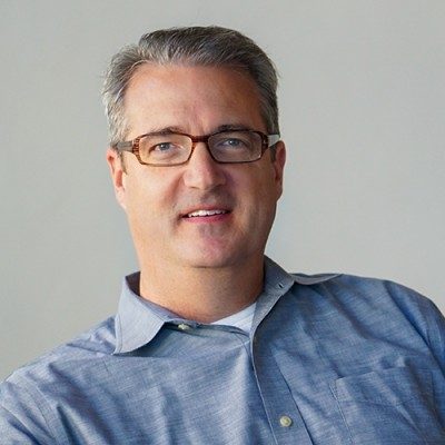 Middle-aged man with gray hair and glasses wearing a blue button-up shirt against a plain background