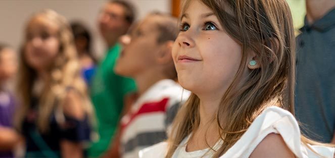 Young girl looking up, surrounded by other children in a blurred background, possibly in a group.