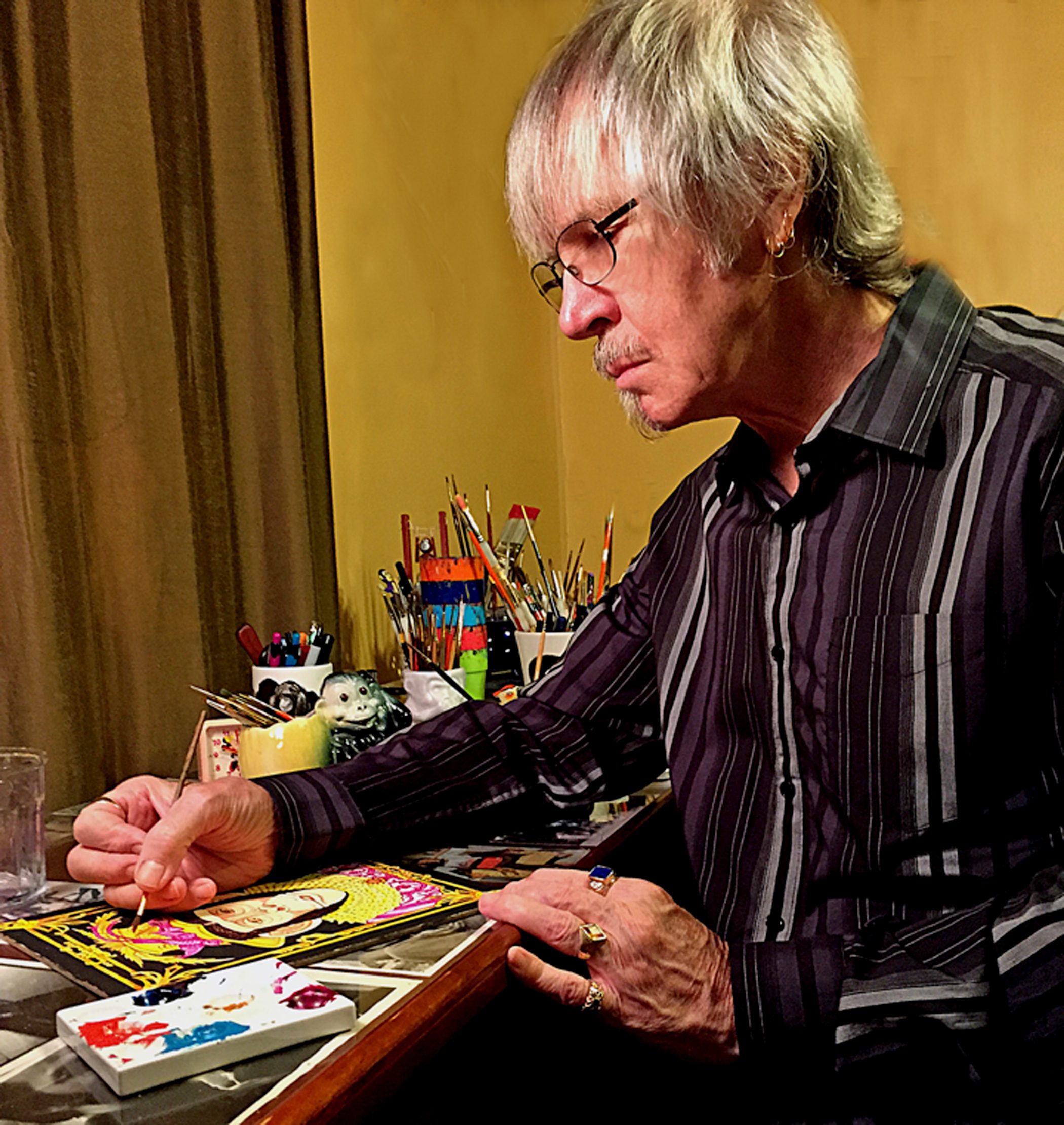Person with glasses paints artwork at a desk filled with art supplies and brushes.