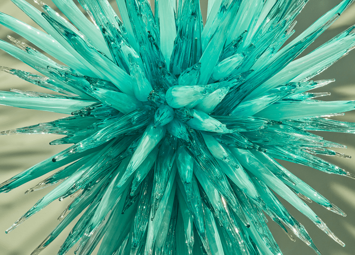 Close-up of teal glass sculpture with pointed, icicle-like structures radiating from a center.